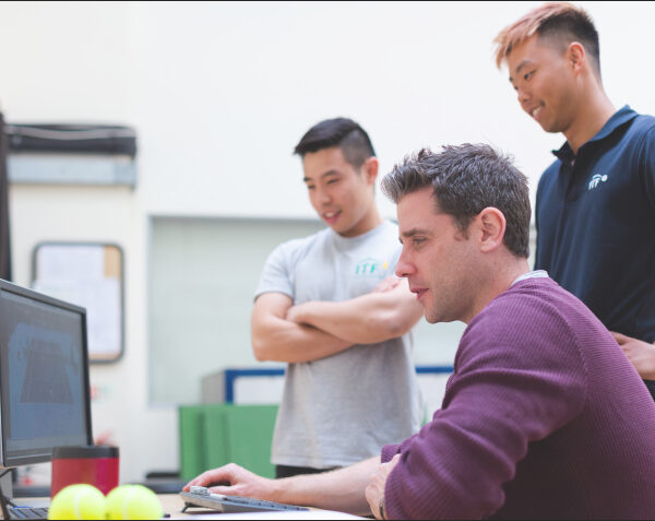 Two men watching another man work at a computer.